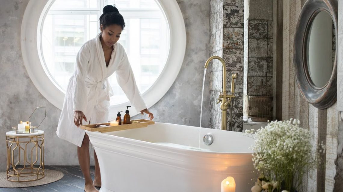 Full body of barefoot African America female in bathrobe putting wooden tray with spa supplies on bath during spa procedure