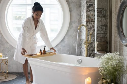 Full body of barefoot African America female in bathrobe putting wooden tray with spa supplies on bath during spa procedure