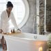 Full body of barefoot African America female in bathrobe putting wooden tray with spa supplies on bath during spa procedure